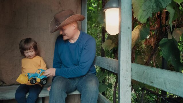 Father cowboy with a small child is sitting on the porch of an old house and playing with his cute baby in toy car. The concept of family pastime and the relationship between children and parents