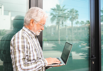 Smiling elderly businessman holding laptop computer on his hands standing out of his office. Mature attractive man wearing eyeglasses using wireless technology