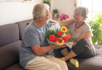 Senior bearded man giving a flower bouquet to his wife celebrating march 8, woman day. Elderly couple looking into each other eyes with love sitting on sofa at home