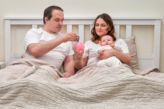 Mother And Father With Baby Boy With Money For Upbringing, Financial Problems Of Parents With Infant Child. Man And Woman Consider Family Budget With Newborn Baby In Bedroom