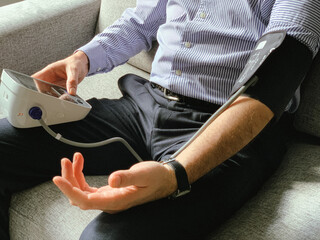 Man is checking blood pressure at home. Sitting on a grey sofa. Monitoring of Seasonal health issues or stress at work Hypertension or hypertension seasonal disorder and problems Taking care of health