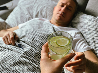 Middle aged man is in bed having flu covid or hangover, drinking water with lemon lime. Refreshing. Holding medical pills in hand. Female hand passing glass of water with lemon lime Care looking after