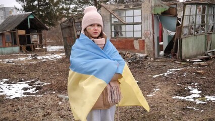 A girl wearing in the Ukrainian flag stands sad against the background of destroyed houses. War in Ukraine