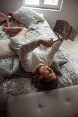 young curly brunette girl in white sweater is lying with gift in hands  on the bed with pillows and smiling happy on the white wall background with window. christmas concept, free space