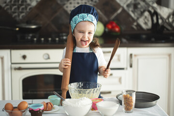 A cute little girl in an apron and a Chef's hat is holding a rolling pin and a wooden spatula,...