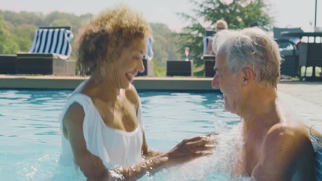 Senior man on summer vacation relaxing by side of outdoor swimming pool being joined by woman who splashed him with water - shot in slow motion