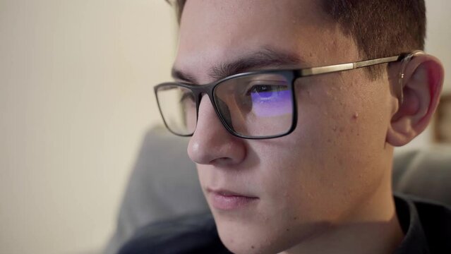 The laptop monitor reflected on the glasses of a young man. Close-up of a focused young businessman. The guy wears computer glasses to reduce eye strain.