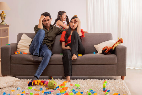 Worried parent sitting on sofa with hands on head and daughter playing elsewhere