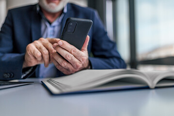 Faceless senior man holding smartphone checking messages and emails while sitting at modern bright office. Focus on hand and phone.