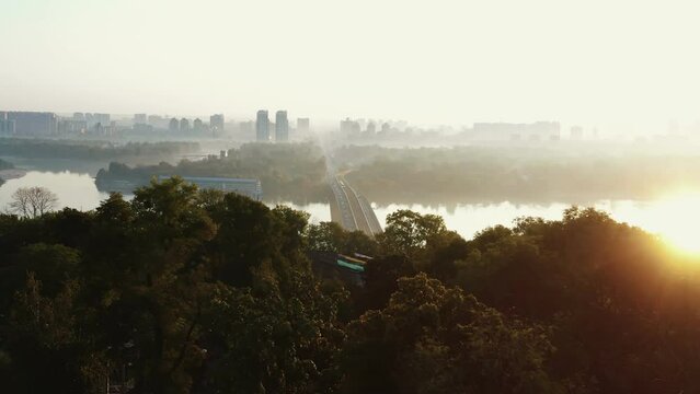 Drone flying over summer trees to reveal amazing bridge with cars, train over Dnepr river, idyllic summer sunrise Kyiv.