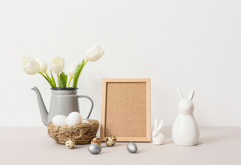 Blank photo frame, nest with Easter eggs, rabbits and tulips on table near white wall