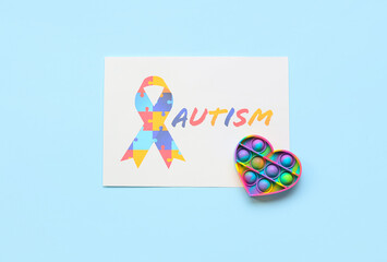 Paper sheet with word AUTISM, awareness ribbon and pop it fidget toy on blue background - obrazy, fototapety, plakaty