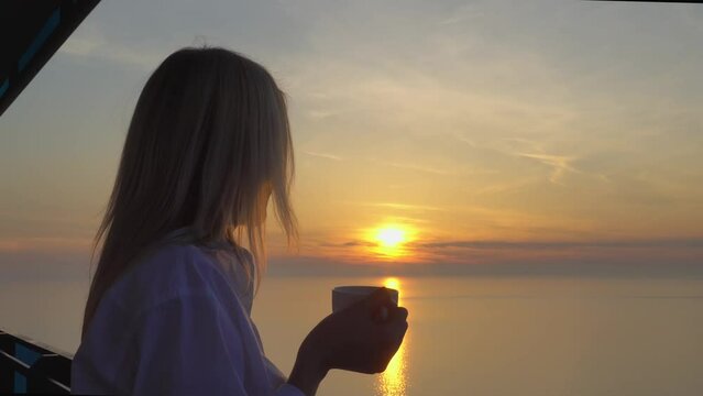 a young beautiful slender blonde woman in a white shirt, drinking coffee or tea while admiring the sunset on the balcony of the upper floor of a multi-storey skyscraper. The setting sun over the sea.