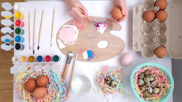 Preparation for Easter. Children's hands mix red and white paint with a brush to get a pink pastel shade. The girl is holding an egg for coloring. Around are eggs, paints and colorful tinsel. Festive