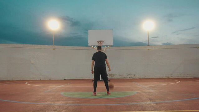 Adult basketball player training on outdoor court during dusk - Dribbling and throwing ball in basket - rear view