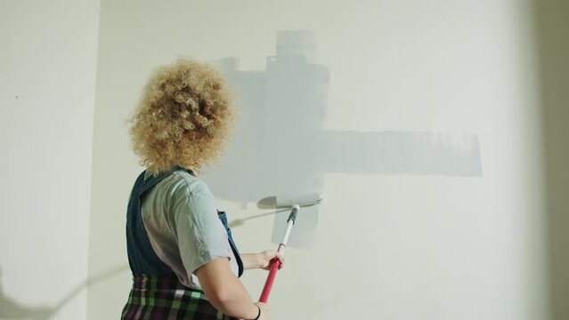Curly hair woman is painting wall in gray using paint rollers. Woman renovation home
