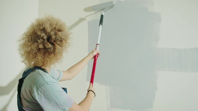 Curly hair Woman in dungarees is painting wall in gray using paint rollers. View from behind, right screen space