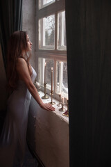 Obraz premium young brunette girl with long hair in blue silk dress is standing in profile near huge white window with old retro candles in vintage style interior background. lifestyle concept, free space