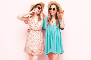 Two young beautiful smiling brunette hipster female in trendy summer dresses. Sexy carefree women posing near pink wall. Positive models having fun. Cheerful and happy. In hats and sunglasses