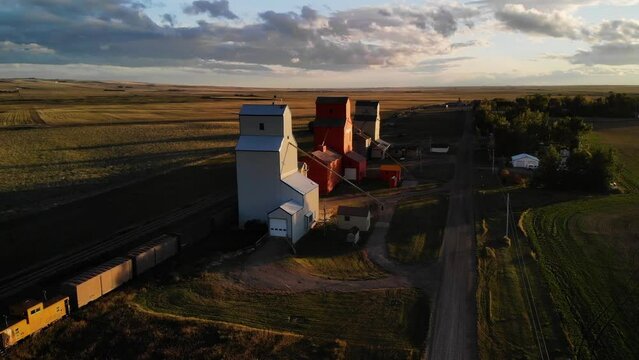 Aerial rotating shot of three Grain Elevators surrounded by agricultural fields on a cloudy day in Alberta, Canada at sunset.