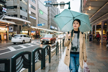 japanese woman pedestrian with camera and plastic bag is walking on sidewalk when it's drizzling. asian female traveler carrying camera is strolling back to hotel after she's done grocery shopping.