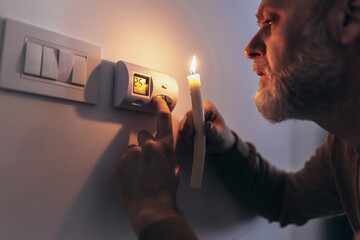 Energy crisis. Man in complete darkness holding a candle to investigate thermostat during a power...