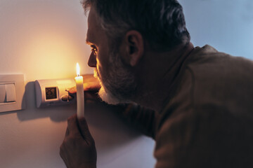 Energy crisis. Man in complete darkness holding a candle to investigate thermostat during a power...