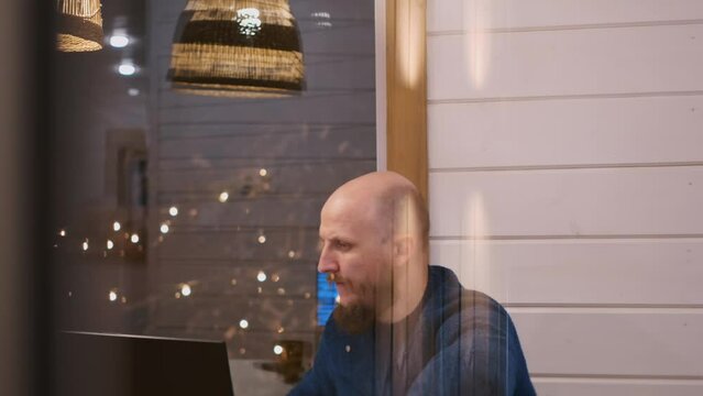 Outside the window, a man with a beard sits at a laptop and does his work in the evening. Remote work at night at home in quarantine.