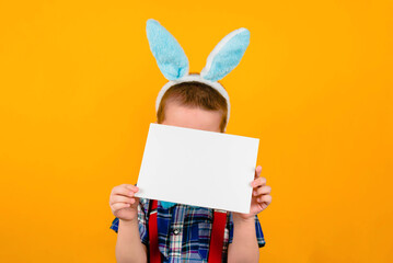 Toddler boy with rabbit ears holds a white piece of advertising paper on a yellow background.