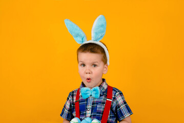 Funny toddler boy with rabbit ears and Easter eggs looks at the camera on a yellow background.