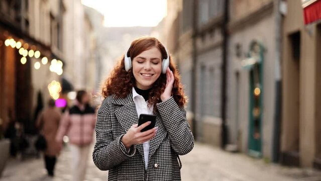 Attractive young red hair woman listening to music in headphone. Slow motion of happy young woman in wireless headphones dancing singing outdoors in city street having fun alone