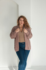 young plus size girl with red curly hair is standing in the beige cardigan and blue jeans and smiling cute on the white wall background. fashion concept, free space