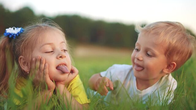 little girl showing her tongue to her brother lying on the grass in the park. happy family kid dream concept. small children play fun in the summer in nature in the park in the green grass