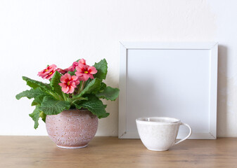 Spring still life. Blank white picture frame mockup on wooden table.Blooming spring pink flower primrose in a pot. Whitewall background. Nature concept.