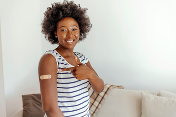 Fototapeta na wymiar illennial pretty African woman show arm with band aid after vaccination in cozy living room. Protection against covid-19 infection, i got vaccinated, be safety. Vaccination, immunizationconcept.
