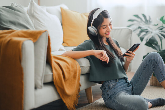 Happy asian woman listening to music from mobile phone while sitting on the rug beside to the sofa at homes, Smiling girl relaxing with headphones in morning, Time to relax.
