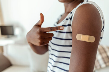 Young African American woman showing her arm with band aid after coronavirus COVID-19 vaccine injection, isolated on gray background. African lady showing vaccinated arm with plaster.