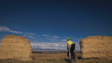 The woman travel on mixed terrain cycle touring with bikepacking. The traveler journey with bicycle bags. Sport bikepacking, bike, sportswear in green black colors. Mountain snow capped, stone arch.