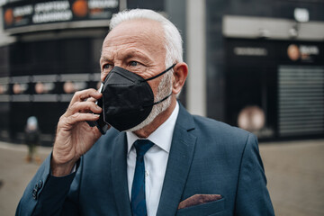 Elegant and positive senior businessman standing on city street and using his smart phone. He is wearing N95 protective face mask as a protection against virus pandemic.