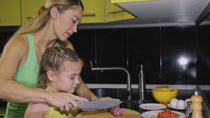 Mother teaching daughter smart girl learning to cook. Young mistress children to cook a Neapolitan egg fried omelette from salame affumicato sausage. Modern Built In Kitchen Appliances.