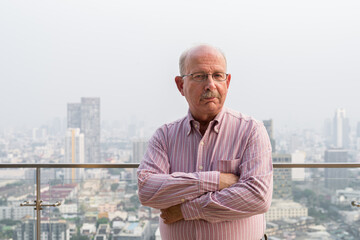 Portrait of senior man looking at camera in city at rooftop with arms crossed