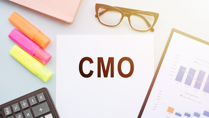 CMO Chief Marketing Officer acronym, written in notepad with glasses, calculator and color markers around on white table