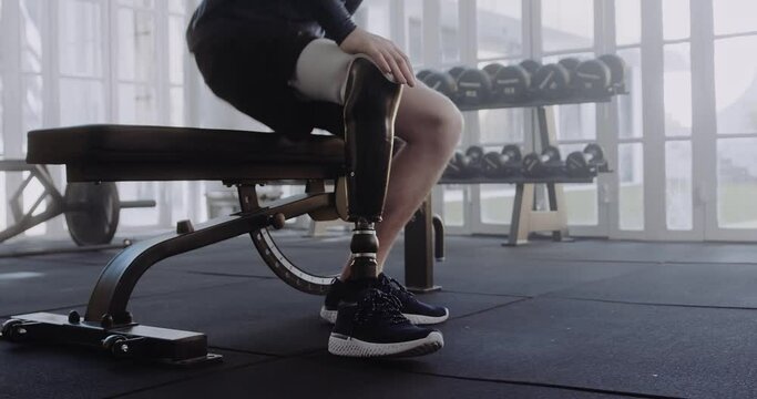 Caucasian male sitting on bench putting on his prosthetic leg. Disabled athlete working out in modern-style gym.
