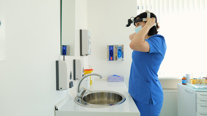 A young woman doctor otolaryngologist puts on a medical mask while standing in her office. Throat and nose treatments. Medicine and health