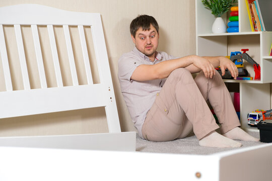 The father is going to put together a children's white wooden bed, sits and looks at the furniture.