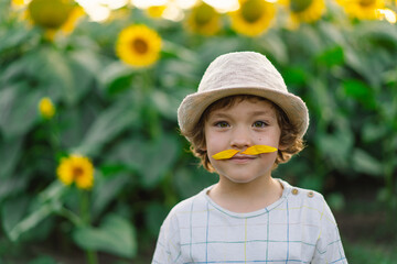 Happy little boy walking in field of sunflowers and making a mustache from sunflower petals. Child...