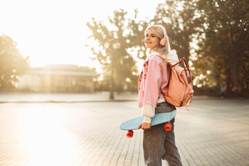 Young cool hipster girl in headphones with skateboard and backpack spends time in the city