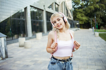 Charming cute young girl listening to music on headphones with a backpack behind her shoulders in the city