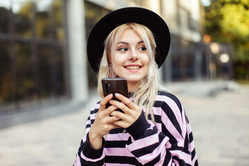 Young charismatic smiling woman in hat using phone outdoors