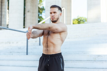 Obraz premium Young male athlete with a naked torso kneads the muscles doing stretching exercise while standing on the stairs outdoors